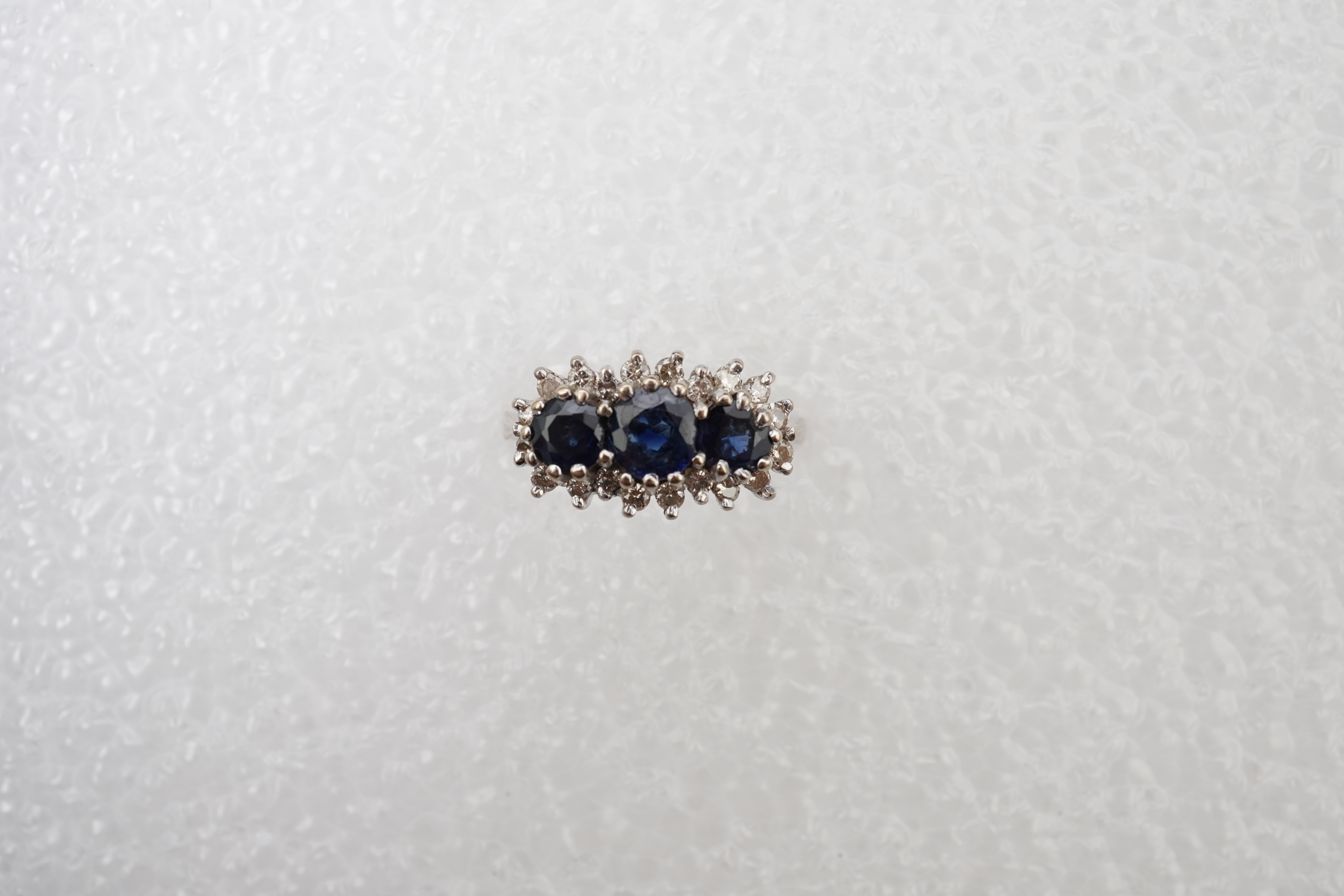 A sapphire and diamond ring, set with three circular-cut and oval sapphires, within a border of brilliant-cut diamonds totalling approximately 0.40 carats, mounted in 18ct white gold, size M1/2, British hallmarks, gross